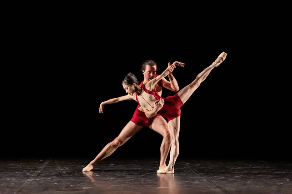 two ballerinas dressed in red leotards are caught in movement on a black empty stage