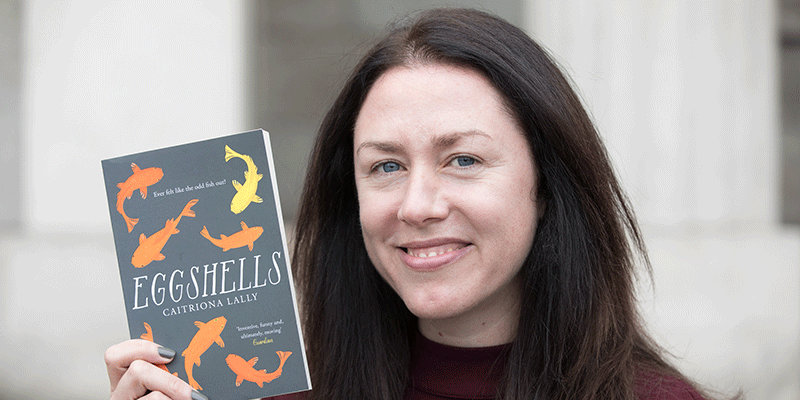 Caitriona Lally, a woman with long, dark hair and blue eyes smiles at the viewer holding her book Eggshells.