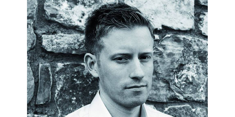 Black and grey headshot of Colin Barrett. The author stands in front of a stone wall and looks away from the viewer