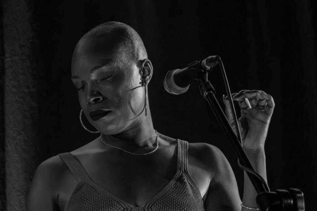 Vocalist Dana Masters stands behind a microphone. She looks down and away from the mic and the viewer with an emotional look on her face. Her hand is raised, ready to curl around the microphone