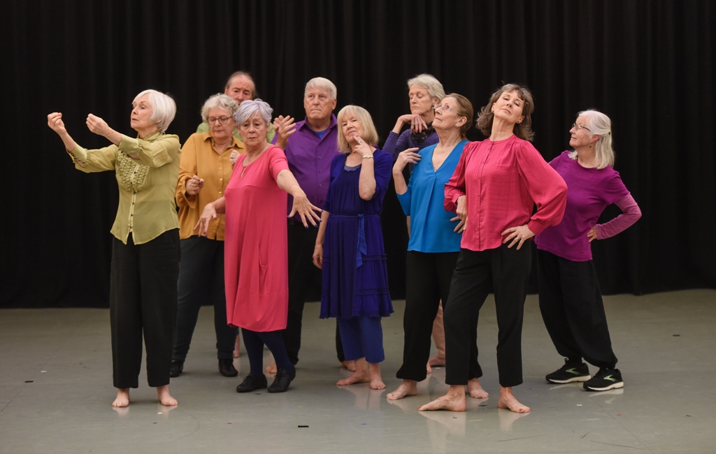 A group of aging men and women are caught in movement on a stage. They wear brightly coloured shirts and dresses and pose in front of a black curtain