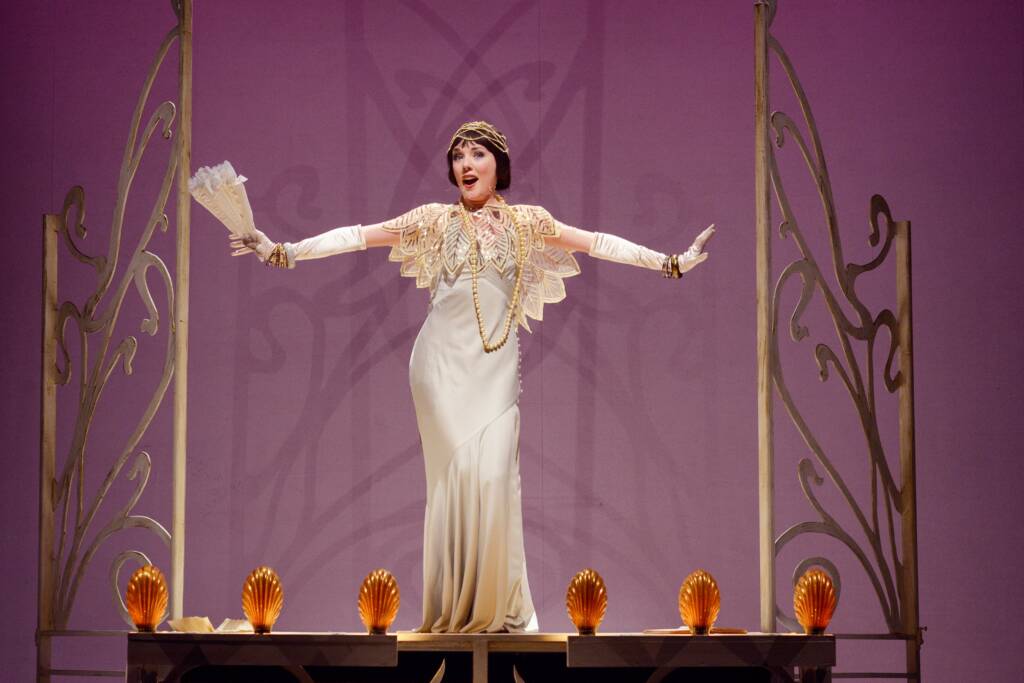 A woman in a flapper dress is cream and gold stands between two ornate art deco fences behind shell shaped orange lights