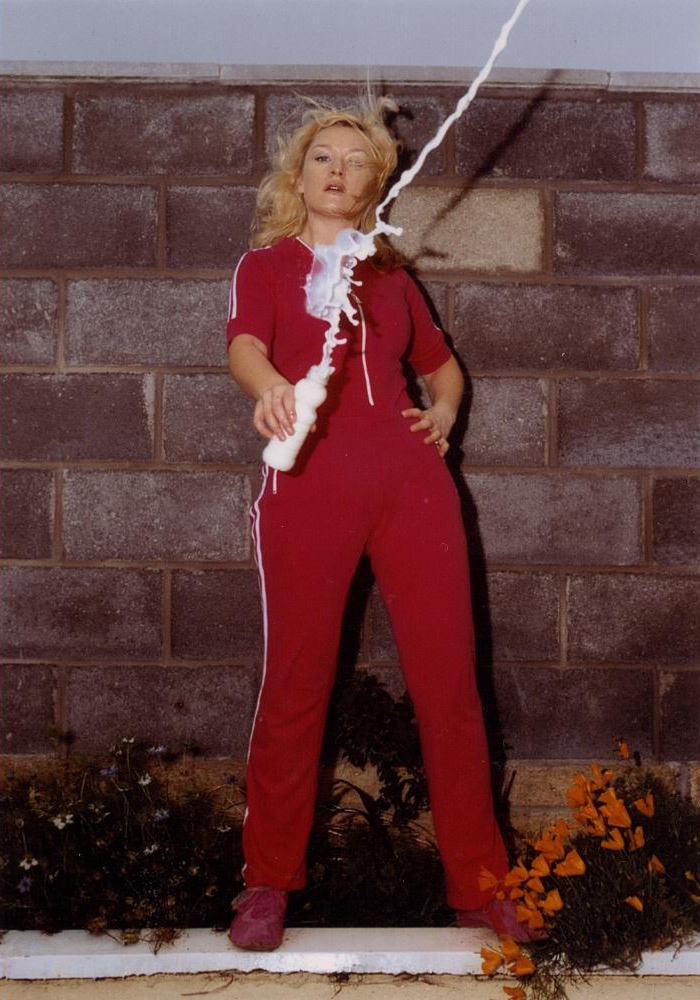A woman stands in a red tracksuit in front of a cinderblock wall. She throws milk from a bottle in front of her which is caught in motion going upward.