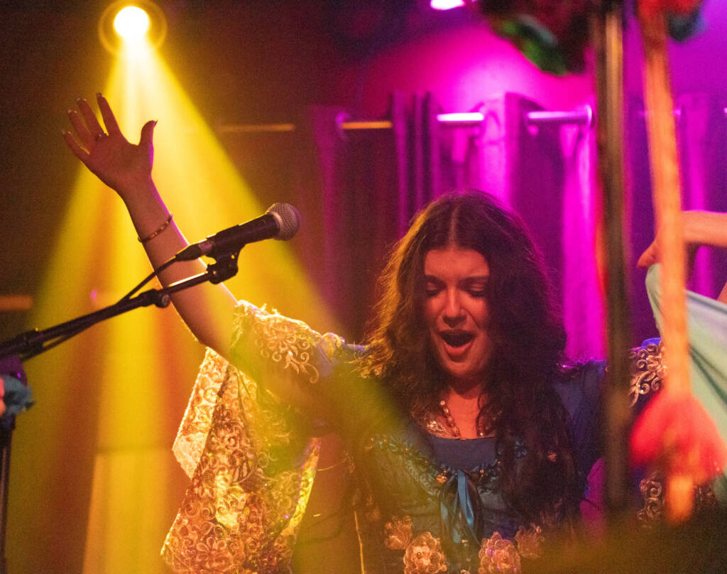 A performer is colourful clothes and long flowing sleeves raises their hands as they sing on stage. They are lit by pink and yellow spotlights