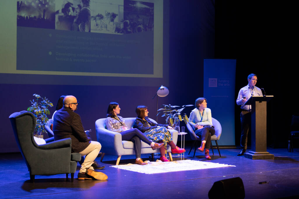 A panel sits and speaks on stage in front of a large screen at Festival Makers Conference 2023