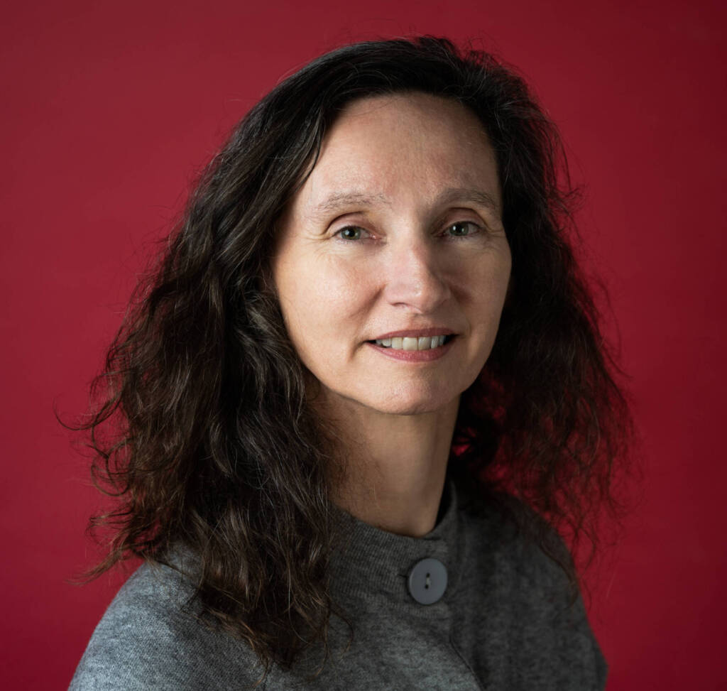 Headshot of Director of the Arts Council Maureen Kennelly. She has shoulder-length dark, wavy hair. She is wearing a grey top with large buttons. The background is a dark red colour.