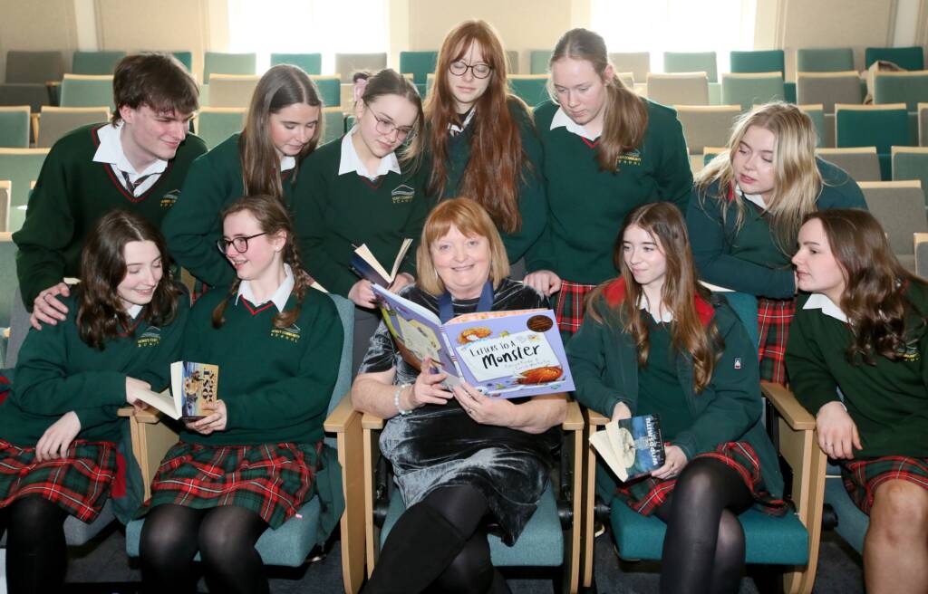 Laureate na nÓg Patricia Forde sits in an auditorium with students in green and red uniforms. They gather as she reads from a book.